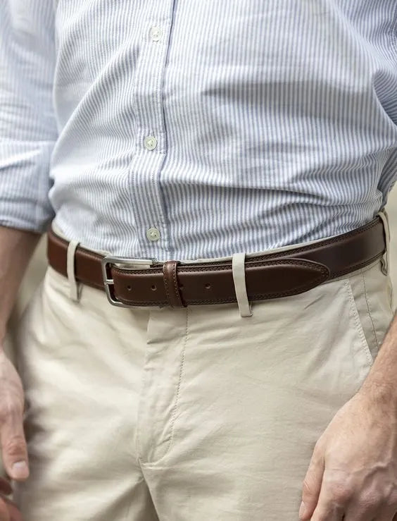 Un homme portant une chemise bleu à rayures fines et un pantalon beige, avec une ceinture en cuir marron. La ceinture est complétée par une boucle métallique, créant une silhouette élégante et soignée