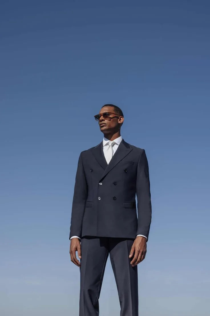 Un homme vêtu d'un costume croisé bleu marine parfaitement ajusté, une chemise blanche et une cravate assortie, portant des lunettes de soleil, pose sous un ciel dégagé, incarnant un style moderne et sophistiqué