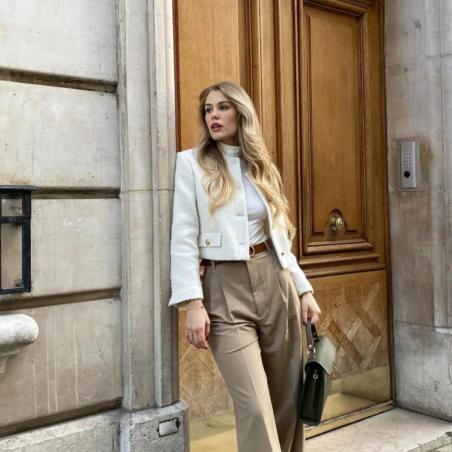 Femme élégante posant devant une porte en bois, vêtue d'un manteau blanc court, d'un pull beige à col montant et d'un pantalon beige ample. Elle porte une ceinture marron et tient un sac à main noir, affichant une allure sophistiquée et moderne.