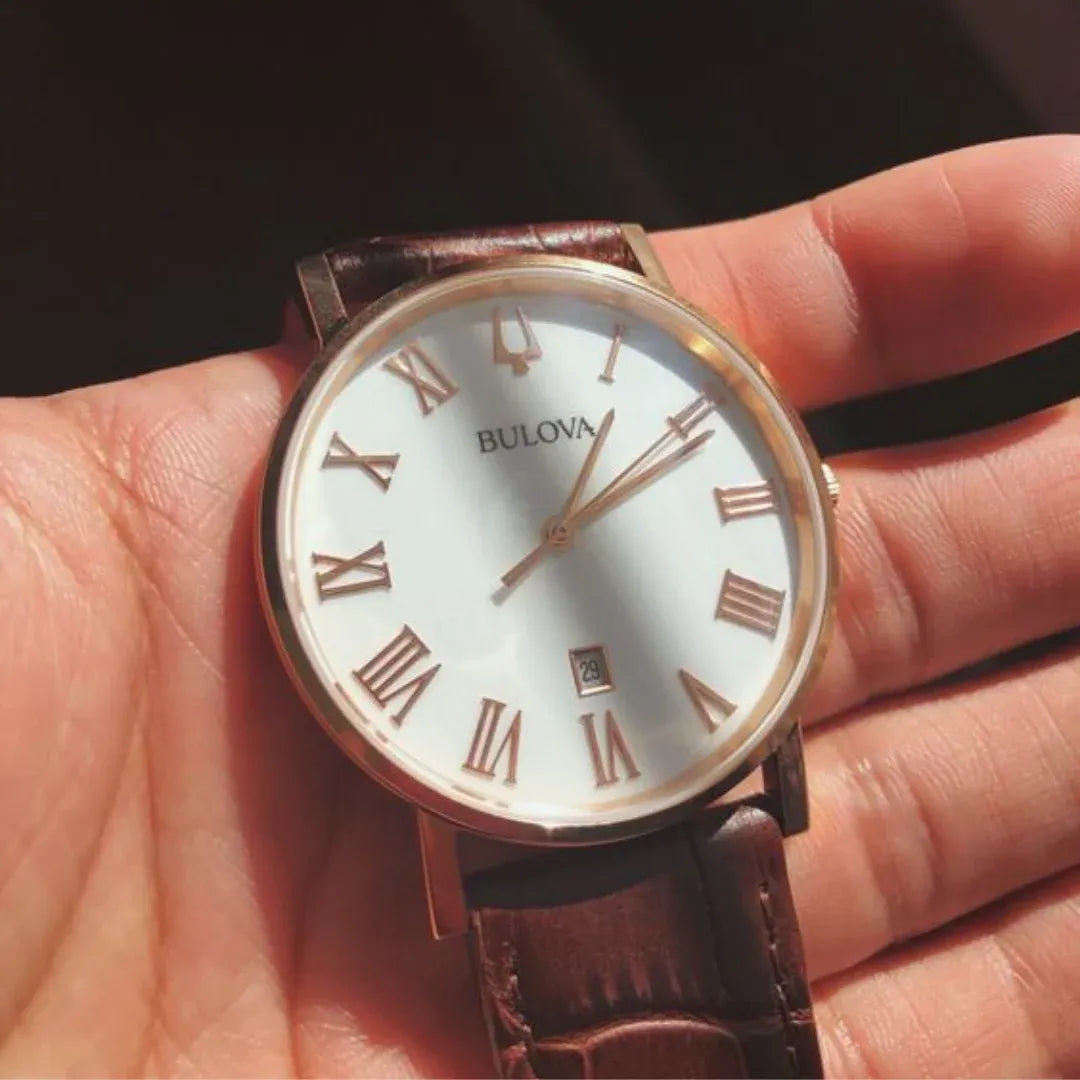 Montre Bulova avec cadran blanc, chiffres romains dorés, aiguilles fines et bracelet en cuir marron. Boîtier en or rose, portée dans une main, mettant en valeur ses détails élégants et sa finition soignée.