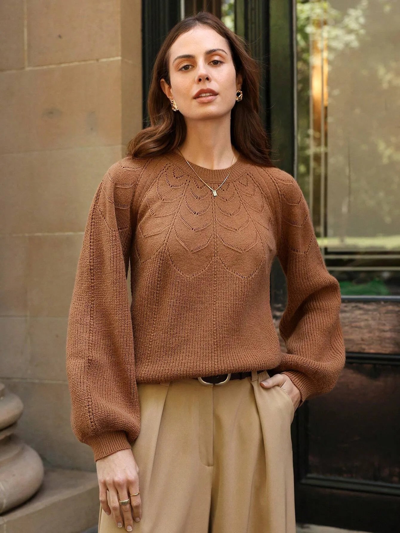 Femme portant un pull marron à manches raglan et un pantalon beige élégant, avec des bijoux dorés et un collier délicat. Elle pose en extérieur, créant une allure chic et décontractée avec une touche de sophistication.