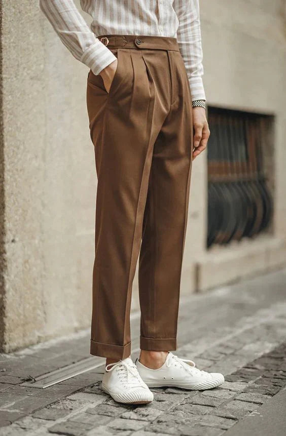 Homme portant un pantalon marron à pinces, une chemise rayée, et des baskets blanches, les mains dans les poches.