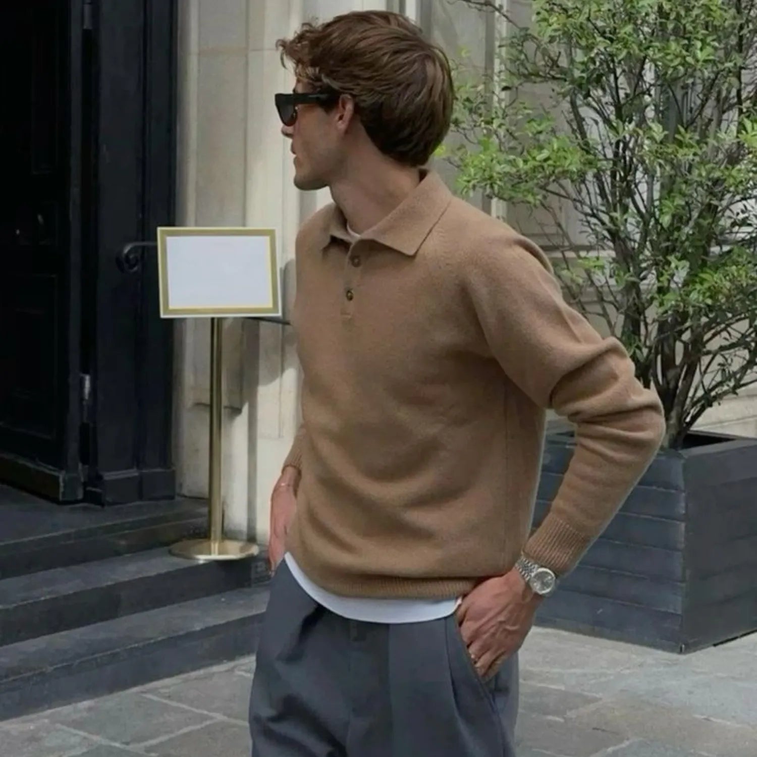 Homme portant un pull beige à col rond et un pantalon marron ample, debout à côté d'un véhicule 4x4. Il affiche un regard sérieux, complétant son look avec des chaussures marron, dans un cadre urbain décontracté et élégant.