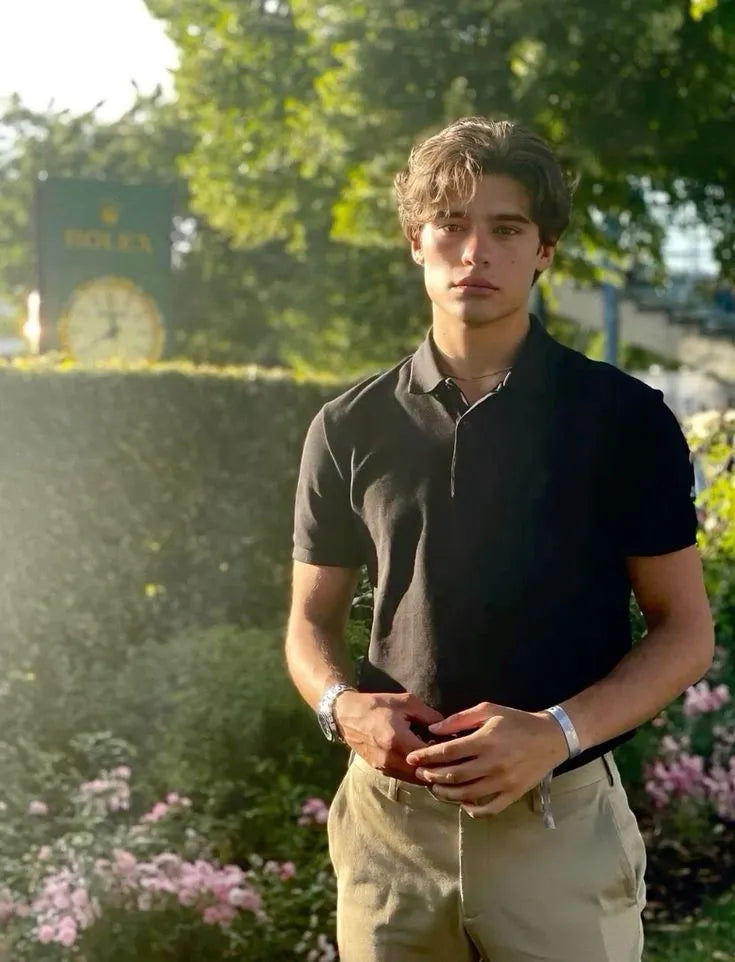 Homme portant un polo noir et un pantalon beige, debout en extérieur avec un regard sérieux. Un bracelet et une montre sont visibles à son poignet, avec une horloge Rolex en arrière-plan, dans un cadre naturel et lumineux.