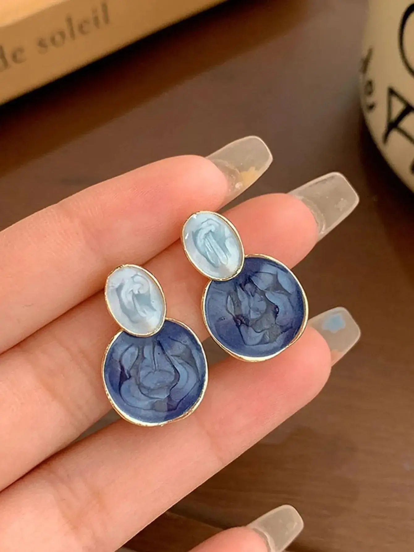 Boucles d'oreilles en alliage métallique doré et résine de qualité, disponibles en bleu clair marbré et bleu marine. Design asymétrique avec cercles marbrés superposés et bordure dorée. Nettoyage à la main conseillé pour préserver leur éclat.