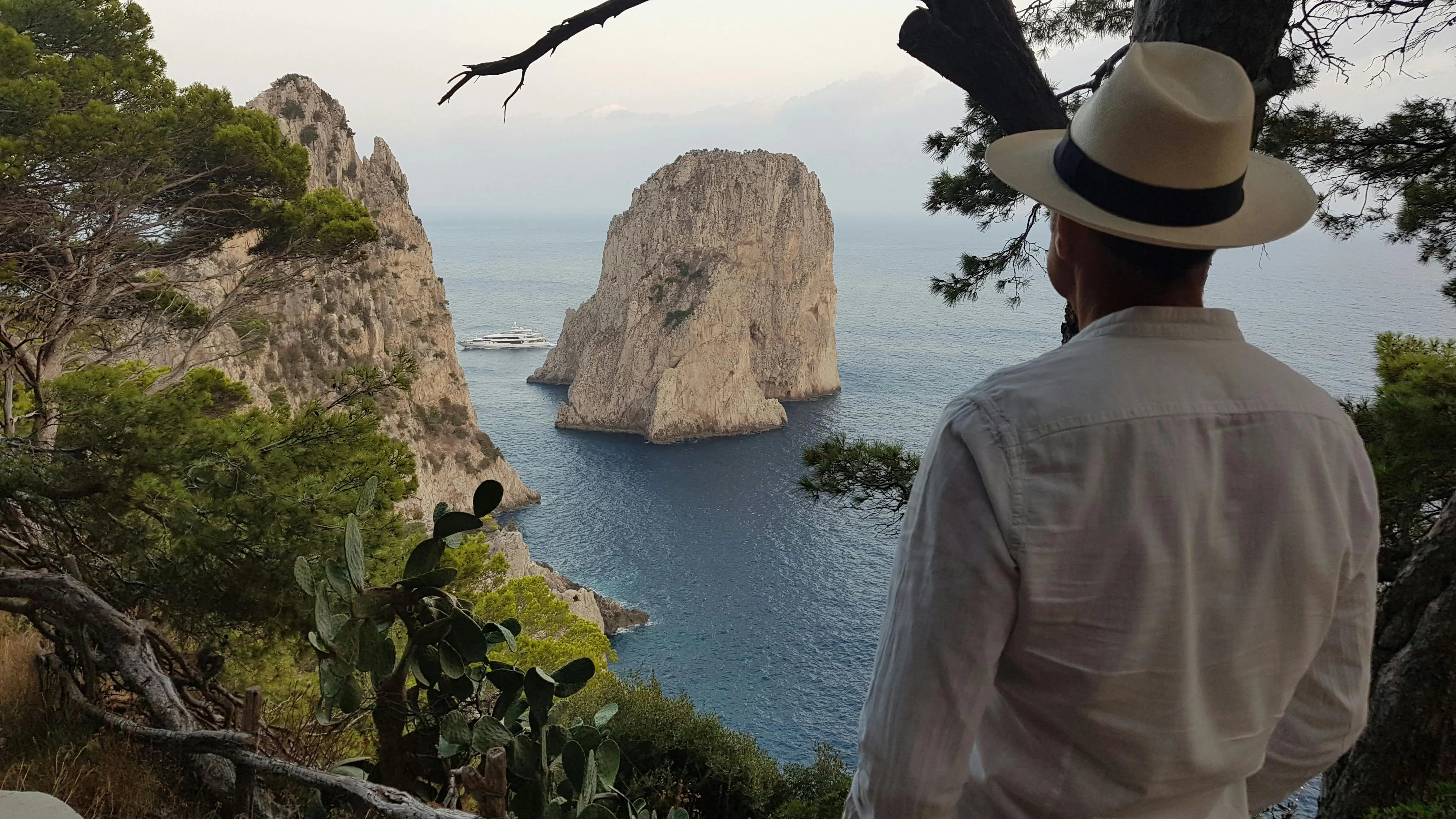 Un homme vêtu d'une chemise blanche et coiffé d'un chapeau Panama regarde la mer depuis un point de vue surplombant des formations rocheuses imposantes et un paysage côtier méditerranéen.