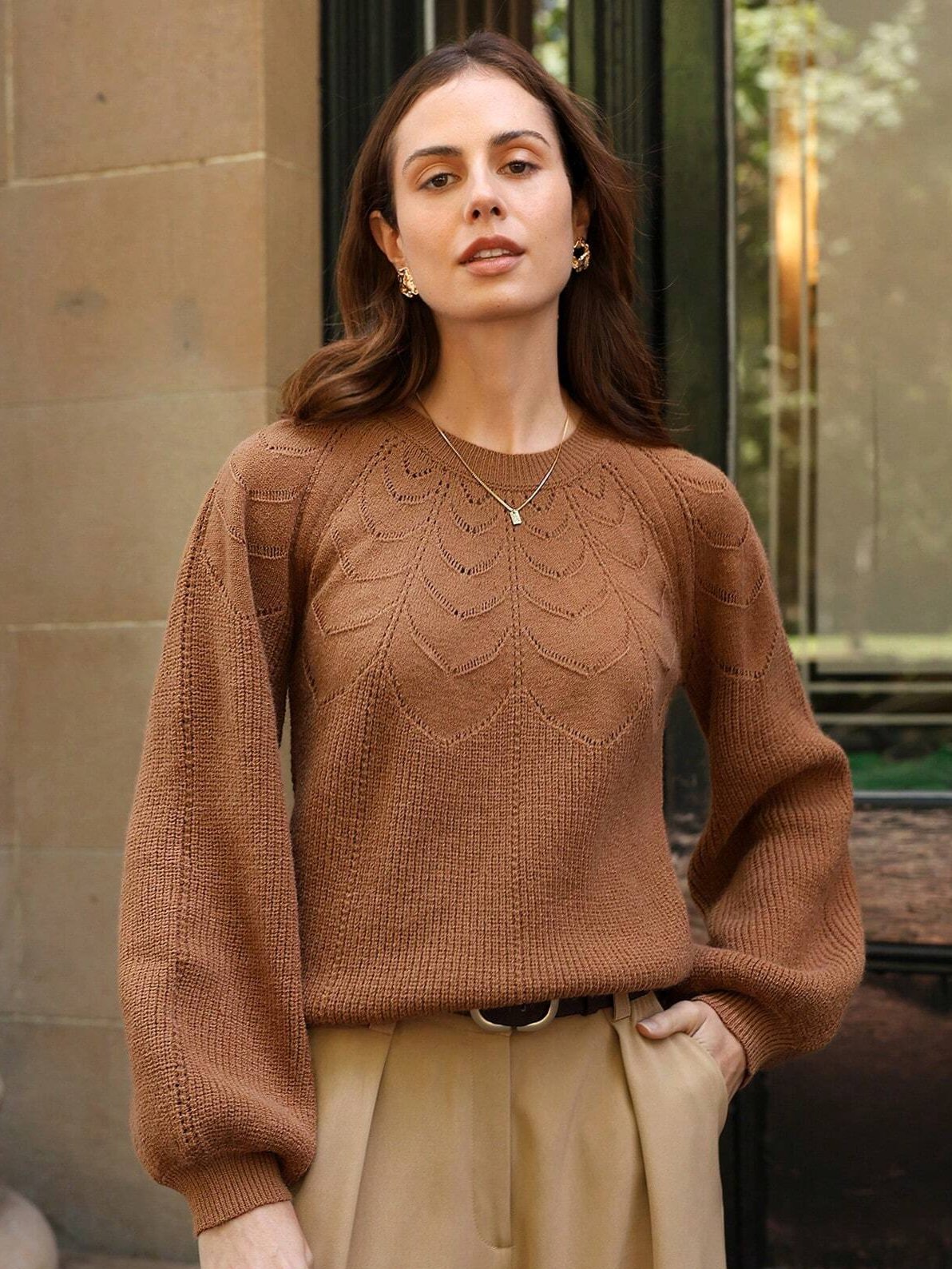 Femme portant un pull en maille avec un motif ajouré dans des tons marron chaud, associant des manches bouffantes et un pantalon beige à taille haute. Elle pose devant une façade en pierre et des fenêtres, créant une ambiance chic et décontractée.